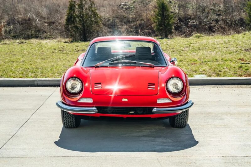 Tampilan depan mobil sport Ferrari Dino 246 GT klasik yang diparkir di permukaan beton, dengan rumput dan pohon di latar belakang, siap untuk momennya di lelang Sotheby Motorsport.