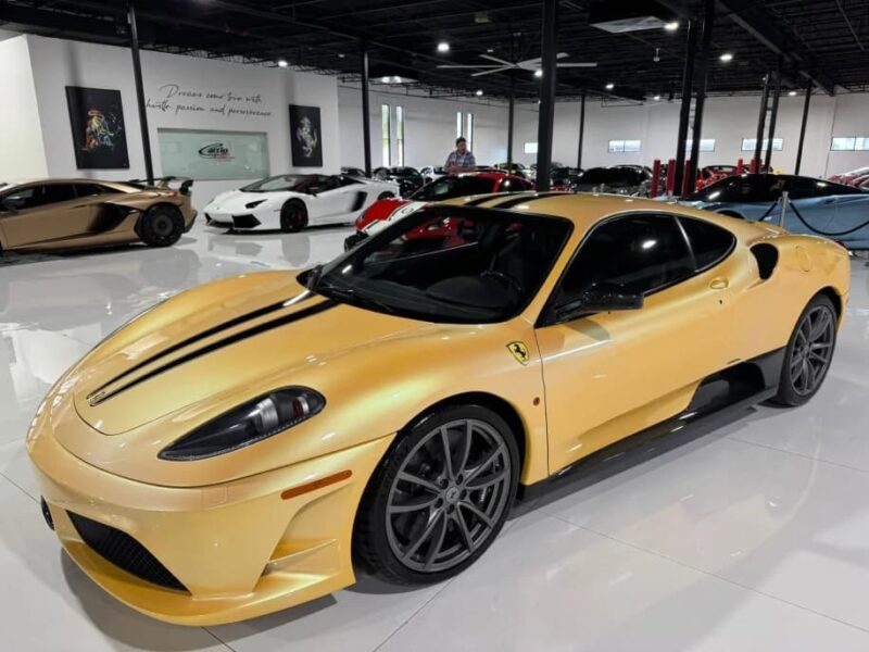 A gold Ferrari 430 Scuderia with black racing stripes is parked in a glossy, well-lit indoor showroom alongside other luxury cars, its Prancing Horse emblem gleaming—an exclusive masterpiece for sale.