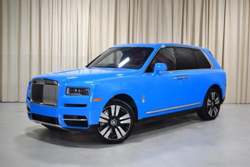 A bright blue Rolls-Royce Cullinan luxury SUV is parked indoors on a light gray floor in front of beige curtains, showcasing the pinnacle of elegance and craftsmanship.