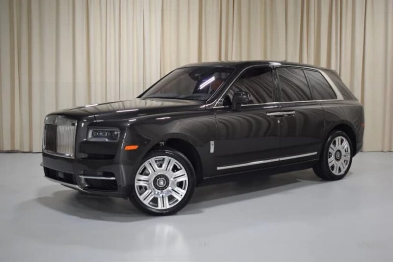 A black Rolls-Royce Cullinan luxury SUV is parked indoors in front of beige curtains under bright lighting.