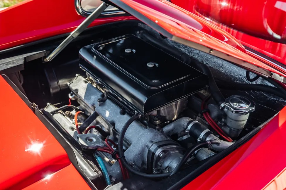 Pemandangan close-up dari mesin Ferrari Dino 246 GT di dalam kendaraan merah dengan tudung terbuka, mengungkapkan berbagai komponen dan kabel, menangkap esensi lineup lelang bergengsi Sotheby Motorsport.