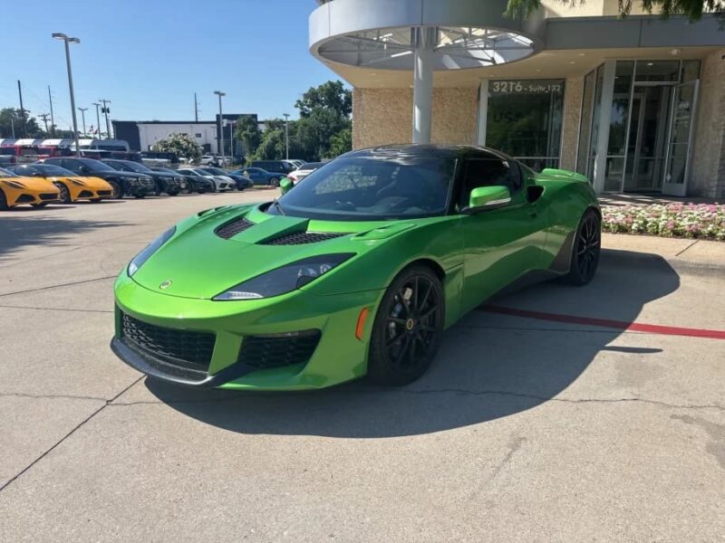 A green sports car with black accents, one of the best green supercars, is parked outside a dealership on a sunny day.