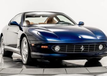 A blue Ferrari coupe, one of the best 2000s cars, is parked indoors on a rotating display platform, with its front and driver's side visible.