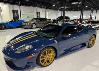 A stunning blue Ferrari 430 Scuderia from the 2000s, with gold racing stripes and gold rims, is parked indoors among other luxury cars. This exceptional Ferrari for sale is a true head-turner.