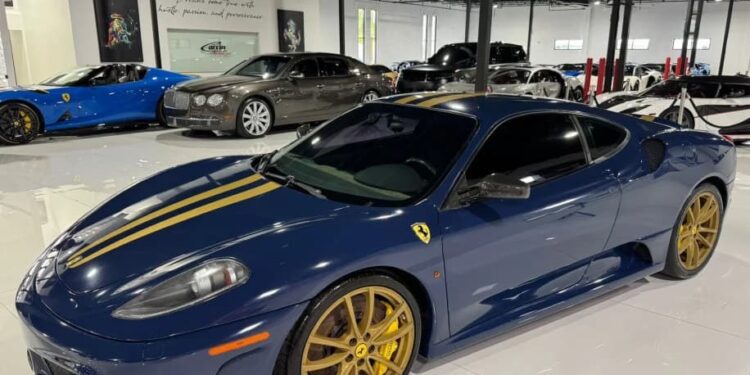 A stunning blue Ferrari 430 Scuderia from the 2000s, with gold racing stripes and gold rims, is parked indoors among other luxury cars. This exceptional Ferrari for sale is a true head-turner.