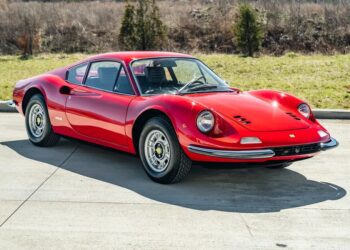 A classic Ferrari Dino 246 GT is elegantly parked on a concrete surface, framed by lush grass and towering trees in the background, hinting at its storied past with Sotheby Motorsport and its allure at auction events.