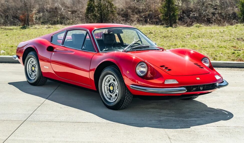 A classic Ferrari Dino 246 GT is elegantly parked on a concrete surface, framed by lush grass and towering trees in the background, hinting at its storied past with Sotheby Motorsport and its allure at auction events.