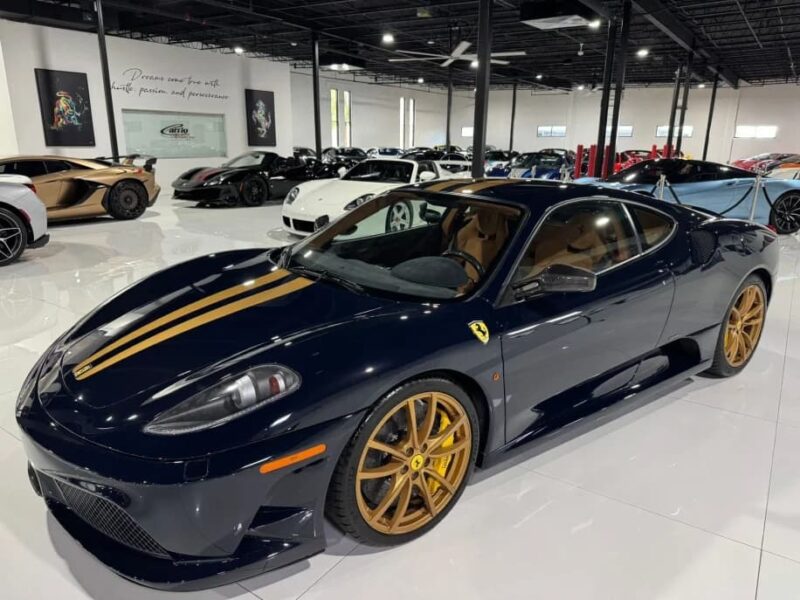 A dark blue Ferrari 430 Scuderia with gold racing stripes and gold wheels is parked in a showroom, surrounded by other luxury cars—an exceptional Prancing Horse for sale.
