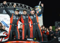 Three race car drivers in matching suits celebrate their Porsche victory on the podium, arms raised high. The iconic race car and cheering team members form a jubilant backdrop after the intense 12 Hours of Sebring spectacle.