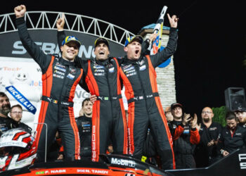Three race car drivers in matching suits celebrate their Porsche victory on the podium, arms raised high. The iconic race car and cheering team members form a jubilant backdrop after the intense 12 Hours of Sebring spectacle.