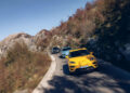 Three cars navigate one of Europe's Best Driving Roads, a narrow, winding mountain path with rocky terrain and autumn foliage. Leading the pack is a yellow car, followed by a blue and gray vehicle, all gliding under a clear blue sky.