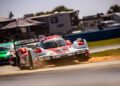 A red and white Porsche race car roars down the track at the 12 Hours Of Sebring, kicking up dirt as a green and white car closely follows. Spectators in the background eagerly anticipate a thrilling battle for victory.