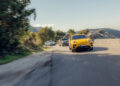 A yellow Porsche car leads two others on a curved mountain road, showcasing Europe's Best Driving Roads with trees and rocky terrain in the background.