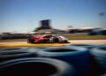 A sleek Porsche race car speeds around the track at the 12 Hours Of Sebring, with stacked tires lining the foreground and blurry buildings in the background under a clear blue sky, chasing victory with every turn.