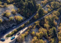 An aerial view reveals a winding European driving road, dotted with several cars—perhaps a Porsche Macan Turbo Electric—masterfully navigating its sharp curves, enveloped by dense trees and rugged rocky terrain.