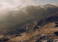 Aerial view of a mountainous landscape with Europe's Best Driving Roads, where a winding path partly cloaked in mist and clouds unfurls under a cloudy sky. Perfect terrain for the Porsche Macan Turbo Electric to glide seamlessly.