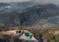 A group of seven cars, including the sleek Porsche Macan Turbo Electric, is parked on a scenic mountain viewpoint overlooking a coastal town and bay. This spot is part of Europe's best driving roads, with stunning mountains and sparkling water in the distance.