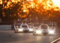 Two Porsche race cars on a track during sunset, with trees in the background and headlights blazing, driven by dreams of victory at the 12 Hours Of Sebring against a warm, glowing sky.