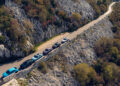 Cars, including a Porsche Macan Turbo Electric, glide along Europe's best driving roads, maneuvering through narrow, winding paths flanked by rugged terrain and sparse trees in a mountainous landscape.