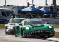 Two race cars speed down the track, with a green Porsche leading the charge. The scene captures the thrill of victory amidst tents and trailers, reminiscent of the 12 Hours Of Sebring.