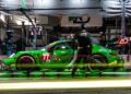 The green Porsche race car number 77 pauses at the pit stop, enveloped by crew members in dark uniforms under bright lighting. Motion blur effects create dynamic lines, capturing the adrenaline of the 12 Hours of Sebring and a taste of imminent victory.
