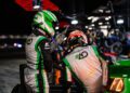 Two race car drivers in matching green and white suits stand by a Porsche during a nighttime pit stop at the 12 Hours of Sebring. One is sitting in the driver's seat while the other points towards the track, envisioning victory ahead.