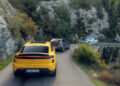 Three cars, led by a yellow one in the foreground, cruise along Europe's Best Driving Roads. The narrow mountain path, enveloped by rocky cliffs and lush greenery, feels tailor-made for a thrill ride in a Porsche Macan Turbo Electric.