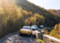 Three cars, with a yellow one leading the way, cruise along a curvy mountain road adorned with the vibrant colors of autumn foliage. Experience this scenic route at its finest in Europe's Best Driving Roads, perhaps behind the wheel of a Porsche Macan Turbo Electric.