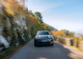 Front view of a sleek, black Porsche Macan Turbo Electric cruising along Europe's Best Driving Roads, where autumn trees and a rocky cliff create a stunning backdrop on a clear day.