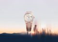 A luxury Rolex wristwatch with a silver and gold metal band is elegantly displayed against a backdrop of mountains and a blurred city skyline at sunset, capturing the essence of Watches & Wonders 2025.