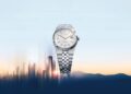 A 2025 Watches silver wristwatch with a patterned face and metallic strap is elegantly displayed against a blurred city skyline and mountain silhouette, set under a pastel sky.