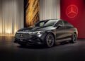 A sleek black Mercedes-Benz CLA is showcased indoors, featuring a modern grille and distinct emblem against illuminated curtains and a red backdrop, highlighting its groundbreaking tech.