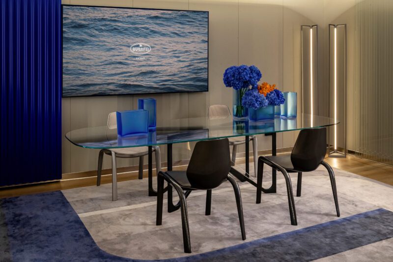 Modern conference room with a glass table, four chairs, and blue and orange flowers. A screen displays a serene water image, while blue and gray décor adds sophistication—a space as sleek as a Bugatti's design.