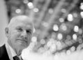 An older man in a suit smiles slightly, standing in front of an out-of-focus background with circular lights—reminiscent of the elegance associated with Bugatti and its legendary W16 engine.