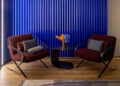 Two maroon chairs with cushions face each other around a small black table with orange and blue flowers, set against a blue corrugated backdrop on a wooden floor, evoking the luxurious design of a Bugatti capsule collection.
