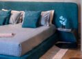 A modern bedroom featuring a blue quilted bed and pillows, alongside a side table displaying blue flowers and a book from the Capsule Collection, exudes an elegance reminiscent of Bugatti's hypercar-inspired design.