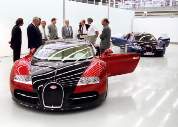 A group of men in business attire stand near two Bugatti Veyron cars, famed for their W16 engines, in a clean, modern showroom with white walls and glossy floors.