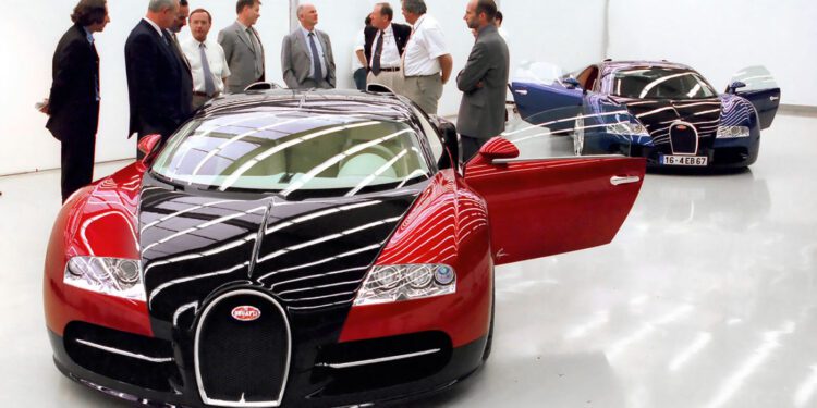 A group of men in business attire stand near two Bugatti Veyron cars, famed for their W16 engines, in a clean, modern showroom with white walls and glossy floors.