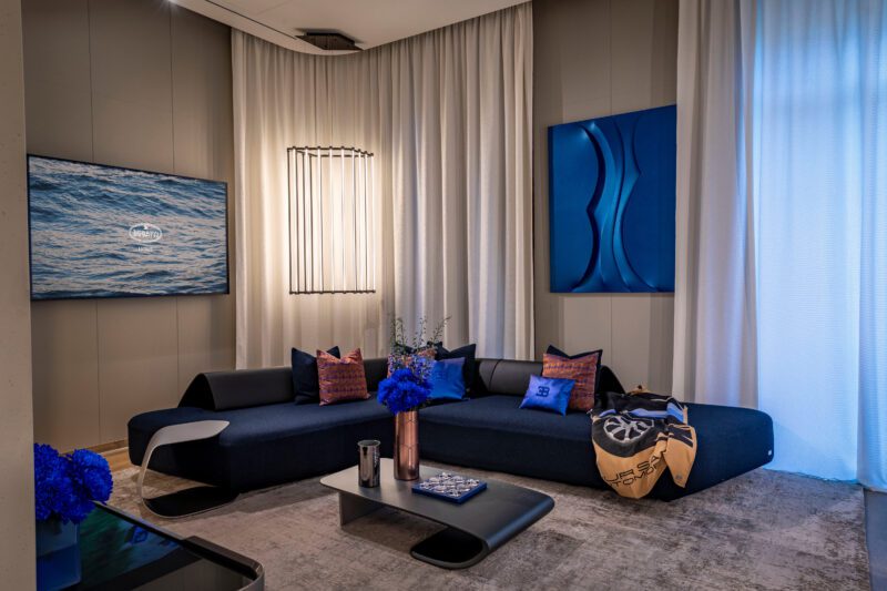 A modern living room features a dark blue sectional sofa, blue artwork, and a flat-screen TV. The minimalist coffee table holds blue flowers and books, reflecting a capsule collection feel. Cream curtains and soft lighting complete this hypercar-inspired space.