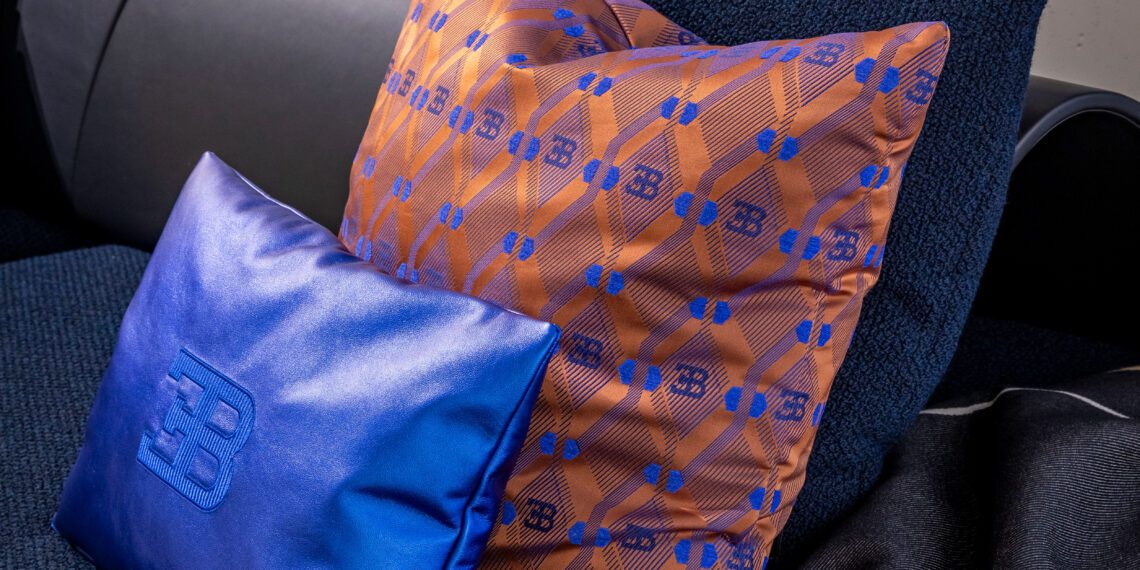 Two decorative pillows on a sofa exude elegance: one is blue with an embossed "B," and the other is orange with a blue geometric pattern, reminiscent of Bugatti's signature style. A perfect touch for any capsule collection, they bring hypercar-inspired sophistication to your living space.
