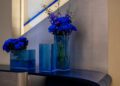 Blue flowers in square blue glass vases are elegantly arranged on a curved countertop, reminiscent of a Bugatti's sleek lines. The minimalist backdrop features a diagonal shadow on the wall, adding an atelier-like touch to this sophisticated home setting.