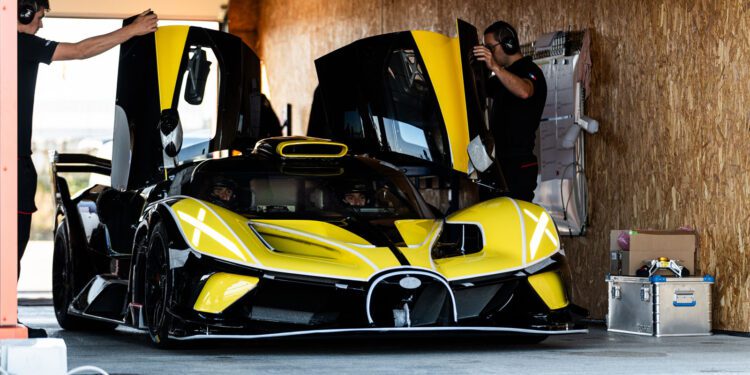 A yellow and black Bugatti Bolide with its butterfly doors open is being prepared by two people in a Miller Motorcars garage.