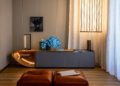 A modern living room features a sleek gray cabinet illuminated by minimalistic lamps. Blue flowers and art decor, reminiscent of a Bugatti hypercar's elegance, adorn the space. A leather ottoman is in the foreground.