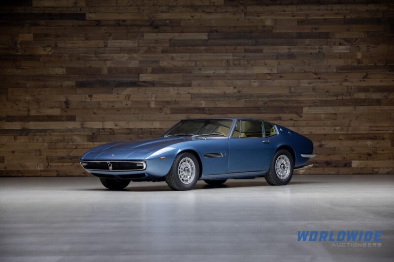 A blue vintage sports car with a sleek design graces the glossy floor, set against a wooden wall background, perfect for an Enthusiast Auction spotlight in Indiana hosted by Worldwide Auctioneers.