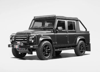 A matte black Land Rover Defender with off-road tires, a front winch, raised suspension, and roof-mounted light bar—this unique creation is photographed in a studio setting, perfect for those seeking custom cars for sale or to showcase in their garage.
