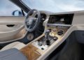 Interior view of a luxury Bentley showcases a leather-trimmed steering wheel, digital dashboard, and wood-accented center console with various controls and gear shift, reminiscent of the sophisticated elegance found in the Continental GT.