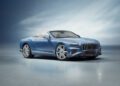 A sleek, blue Bentley convertible gracefully sits on a reflective surface, its design capturing the essence of luxury, set against a blurred blue background.