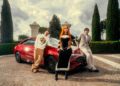 Three people pose in fashionable clothing in front of a red Mercedes-Benz CLA parked on a stone driveway, with tall green trees and a gated estate, channeling an Ice Spice remix vibe.