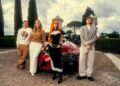 Four people pose in front of a red Mercedes-Benz CLA on a cobblestone driveway, giving off major Ice Spice remix vibes, with tall trees and an ornate gate in the background.