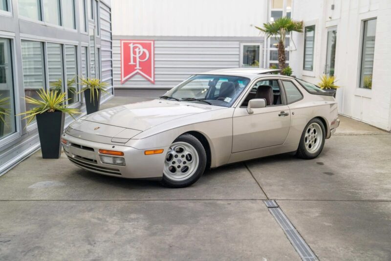 A silver Porsche 944, an unsung classic from the transaxle Porsche era, is parked in an outdoor area near modern buildings and potted plants.